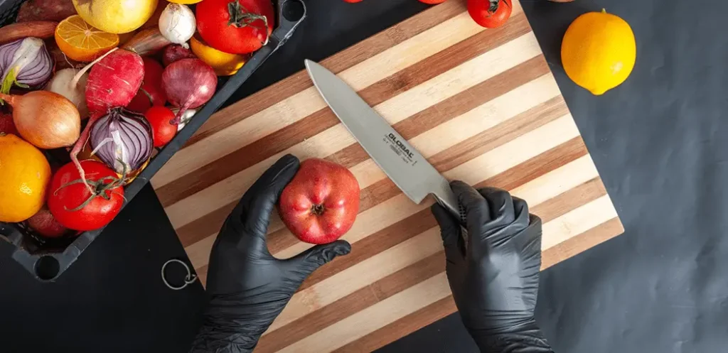 Chef sujetando un cuchillo para chef profesional Global G-2 apunto de cortar una manzana sobre una tabla para picar y verduras al rededor